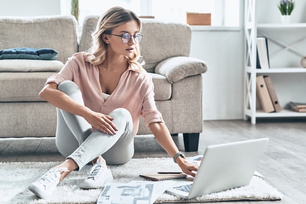 Poner contenido nuevo en su blog. Mujer joven pensativa en gafas trabajando usando la computadora mientras el piso en casa