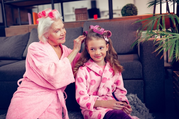 Poner rizadores de pelo. Cuidando a la abuela de pelo gris poniendo rulos en el cabello de su linda niña