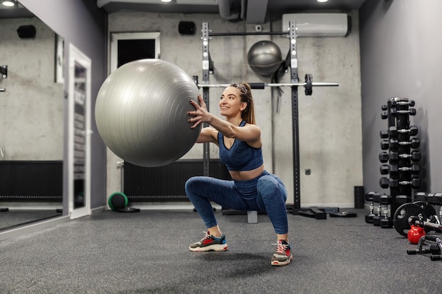 Ponerse en cuclillas con una gran bola gris Una mujer atractiva en ropa deportiva hace ejercicios para quemar los músculos