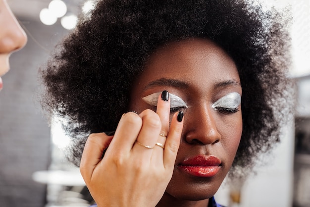 Ponerse sombras de ojos. Joven estilista rubia vistiendo esmalte de uñas negro trabajando con el modelo