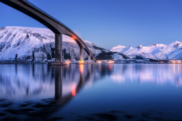 Ponte com iluminação, montanhas cobertas de neve, vila e céu azul com belo reflexo na água