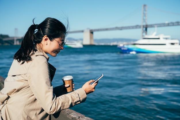 Ponte da Baía de São Francisco Oakland e antena da Ilha do Tesouro em segundo plano. jovem usando smartphone conversando online contra o corrimão de madeira perto do mar. iate navegando no oceano azul.
