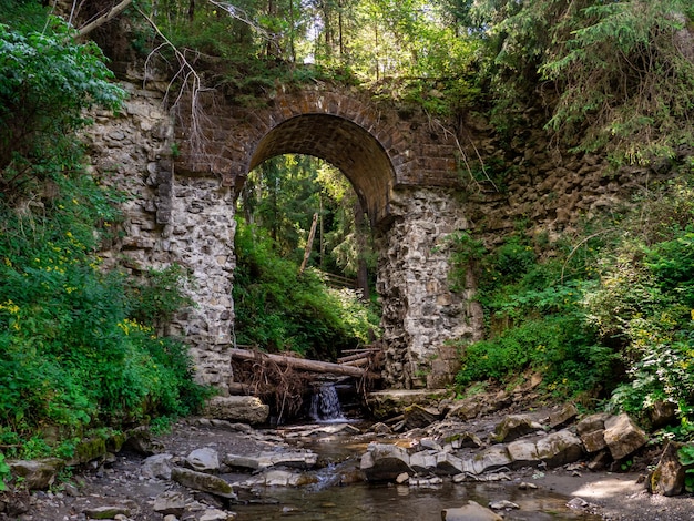 Ponte destruída em arco de pedra velha sobre um pequeno rio numa floresta montanhosa