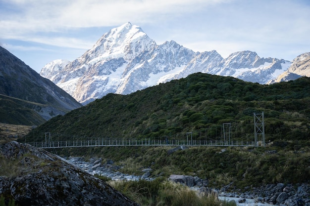 Ponte giratória que conduz através de um vale alpino com uma enorme montanha coberta de neve atrás dela, o Monte Cook, Nova Zelândia