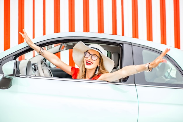 Poortrait einer jungen glücklichen Frau, die während der Sommerferien mit erhobenen Händen auf dem roten Wandhintergrund aus dem Fensterauto zieht