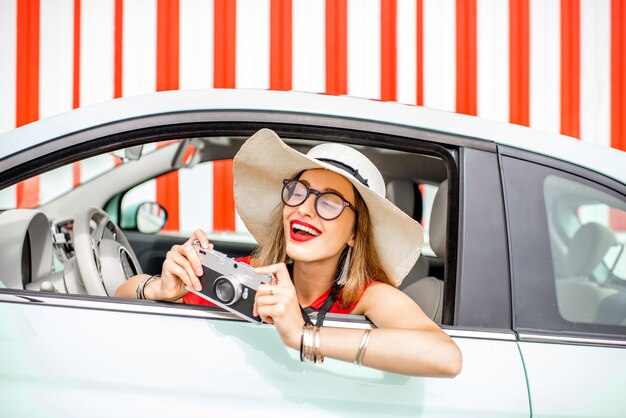 Poortrait de una joven mujer feliz saliendo de la ventanilla del coche con cámara de fotos en el fondo de la pared roja durante las vacaciones de verano