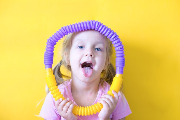 Pop-Röhre. Anti-Stress-Kunststoffspielzeug aus einer Pop-Röhre in den Händen eines Kindes. Fröhliches kleines Mädchen, das mit einer Spielzeugzappel-Popröhre spielt. Trend von 2021. Gelber Hintergrund