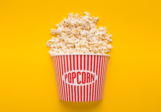 Popcorn im klassischen gestreiften Eimer auf gelbem Hintergrund. Flauschige Maisstreuung aus Papierbox, Kopierraum. Fast Food und Filmsnack, Unterhaltungskonzept, Kopierraum
