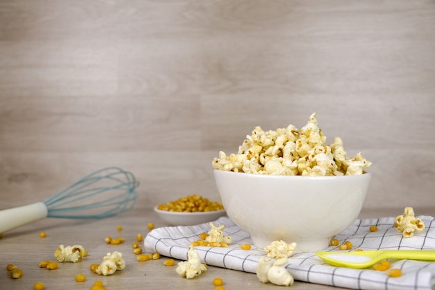 Popcorn in der weißen Schüssel mit Pastelllöffel-, Salz-, Handmischer- und Maissamen auf hölzernem Hintergrund