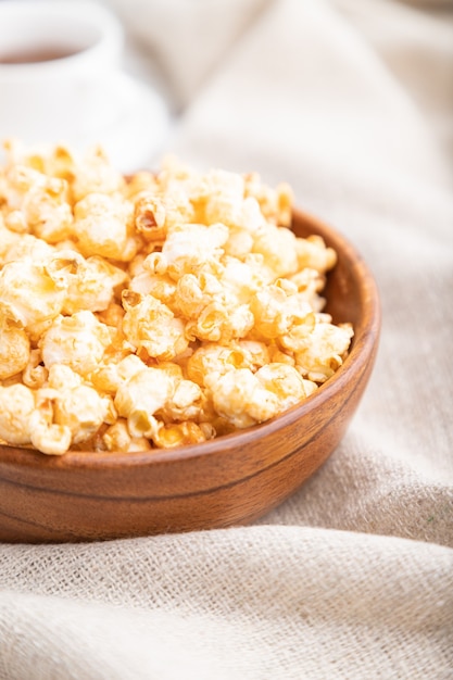 Popcorn mit Karamell in der Holzschale und einer Tasse Kaffee auf weißem hölzernem Hintergrund und Leinentextil. Seitenansicht, Nahaufnahme, selektiver Fokus.