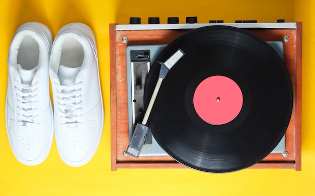 Popkultur. Weiße Hipster-Turnschuhe, Vinyl-Spieler auf gelbem Hintergrund. Retro-Stil. 80er Jahre