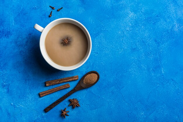 El popular calentamiento asiático bebe té de masala en un Ñ blanco con cilantro, una cuchara de madera con canela y un tallo de rosa sobre un fondo azul clásico.