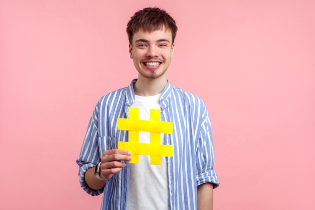 Foto popularidad en las redes sociales. retrato de un joven feliz de cabello castaño con un pantalones casual que sostiene un gran cartel amarillo, sonriendo con entusiasmo. tiro de estudio interior aislado sobre fondo de color rosa