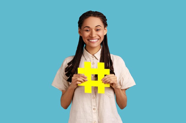 Popularidade na Internet Retrato de mulher feliz com dreadlocks pretos segurando o símbolo de hashtag promovendo tópico viral na rede social marcando tendências de blog Foto de estúdio interior isolada em fundo azul