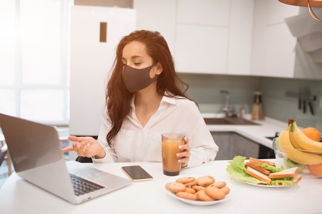 Por causa da pandemia de coronavírus, uma mulher isolada em casa. Ela trabalha em casa, está usando uma máscara e tem uma videoconferência em seu laptop.