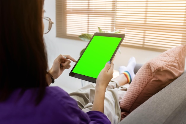 Por cima do ombro garota usando computador tablet no sofá na sala de estar com tela verde em branco Trabalho em casa conceito