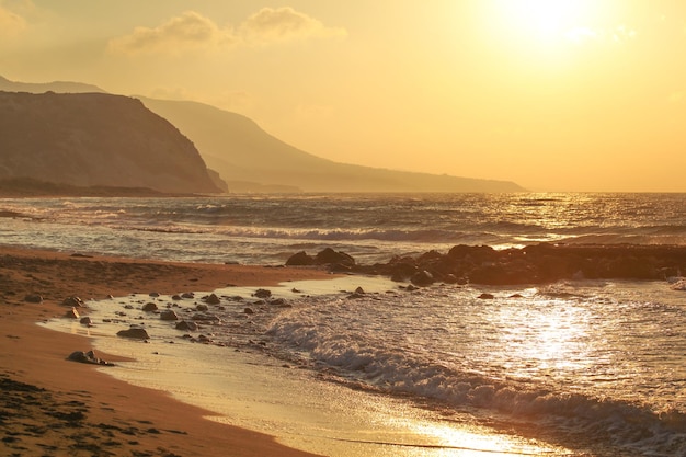 Pôr do sol na praia selvagem vazia. pequenas ondas sobre rocha, colinas no fundo nebuloso.