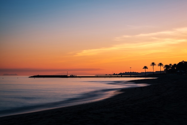 Pôr do sol numa praia da Costa del Sol em Marbella, com a silhueta de um cais e Gibraltar.