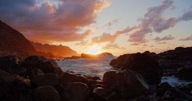 Pôr do sol sobre o oceano Nuvens laranja e silhueta de montanhas de formações rochosas Ondas de praia rochosa espirrando Meditação