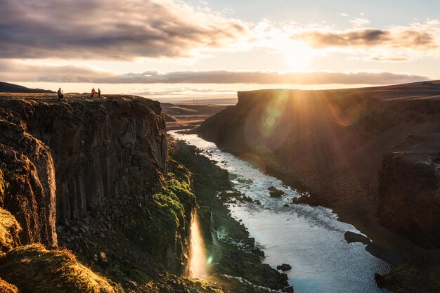 Pôr do sol sobre Sigoldugljufur com alpinista em pé no penhasco e cachoeira fluindo no desfiladeiro nas Terras Altas da Islândia no verão na Islândia