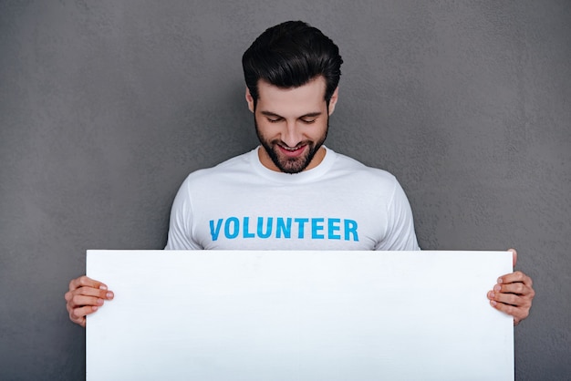 Por favor, ajude a comunidade! jovem confiante em uma camiseta de voluntário segurando um quadro branco e olhando para ele com um sorriso em pé contra um fundo cinza