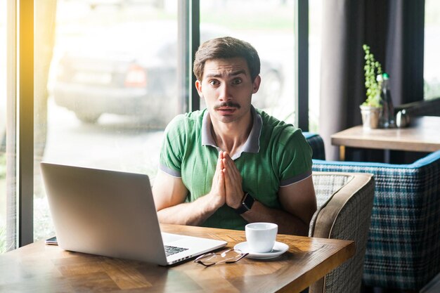 Por favor, perdoe-me ou ajude o jovem empresário preocupado em camiseta verde sentado e olhando para a câmera e implorando para ajudar ou perdoar o conceito de problema de negócios tiro interno perto da grande janela durante o dia