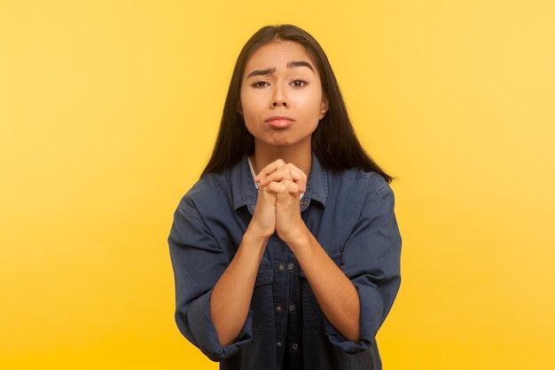 Por favor. Retrato de menina implorando em camisa jeans pedindo cordialmente e de mãos dadas em gesto de oração, implorando por ajuda, pedido sincero. tiro de estúdio interno isolado em fundo amarelo