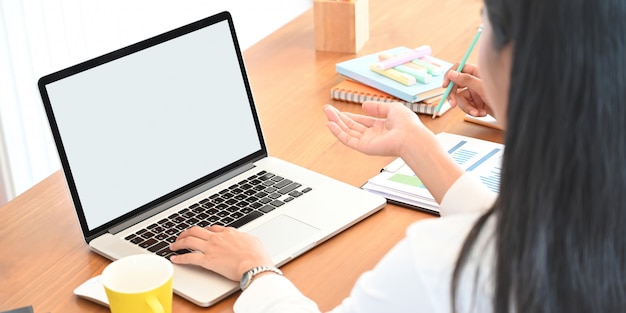 Por trás do tiro da mulher criativa, trabalhando no laptop do computador com tela em branco branca que colocar na mesa de trabalho de madeira e rodeado por equipamento de escritório e estacionário como pano de fundo.