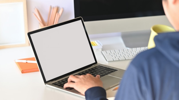 Por trás do tiro do programador inteligente segurando a xícara de café e digitando no laptop de tela em branco branco enquanto está sentado sobre seu monitor de tela preto na mesa de trabalho moderna.