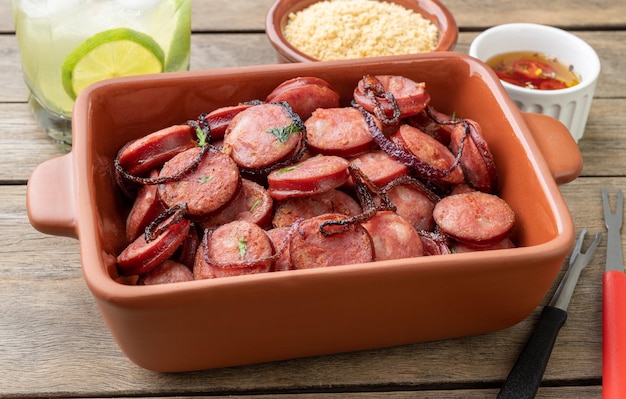 Porção de linguiça calabresa grelhada com farofa de cebola e caipirinha sobre mesa de madeira