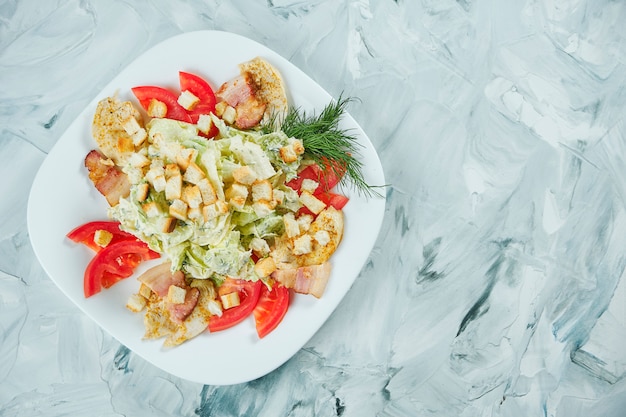 Porción de ensalada César con picatostes, lechuga, salsa blanca y pollo frito en plato blanco sobre mesa gris. Comida de restaurante saludable