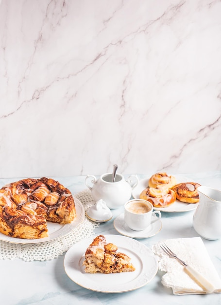 Porción de rollos de canela caseros en una placa blanca sobre una mesa blanca con bollos de café y pastelería