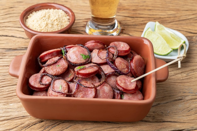 Porción de salchicha calabrese a la parrilla con cebolla, limón, farofa y cerveza sobre una mesa de madera