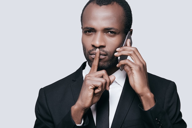 Porfavor guarda silencio. Apuesto joven africano hablando por teléfono y gesticulando mientras está de pie contra el fondo gris