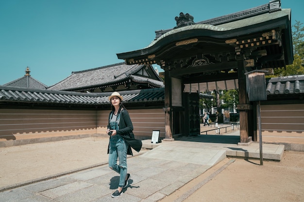 portão de madeira da entrada do templo shitennoji com turista feminina caminhando. comprimento total do jovem viajante deixando o santuário indo para a próxima atração. fotógrafo de mulher carrega câmera em dia ensolarado.