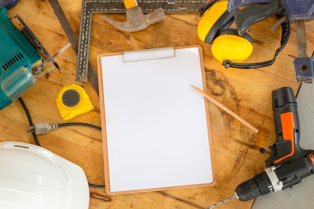 Portapapeles en blanco de maqueta con hoja blanca y lápiz en mesa de carpintero Cascos de seguridad blancos y herramientas de trabajo en banco de trabajo en taller de carpintería