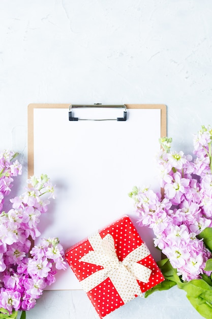 Portapapeles con papel en blanco blanco, caja de regalo presente y flores sobre fondo blanco.