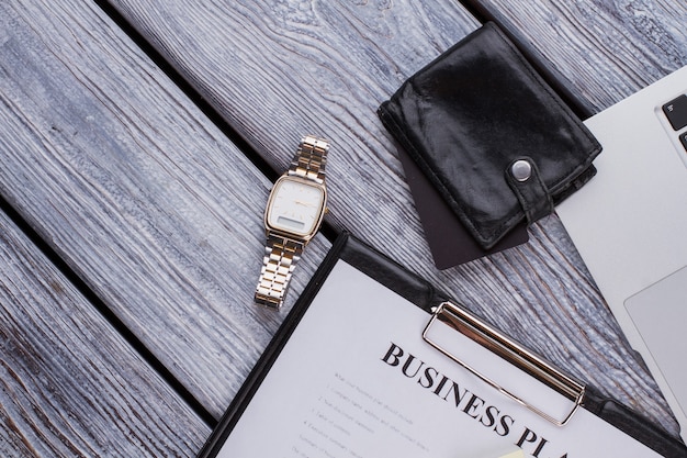 Portapapeles con plan de negocios y reloj de lujo. Accesorios de empresario en una mesa de madera blanca.