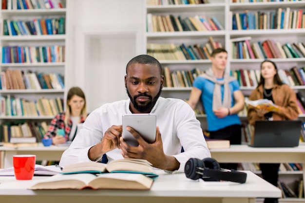 Portarit de jovem concentrado jovem estudante masculino Africano Africano na camisa branca, estudando na biblioteca com seus amigos no espaço