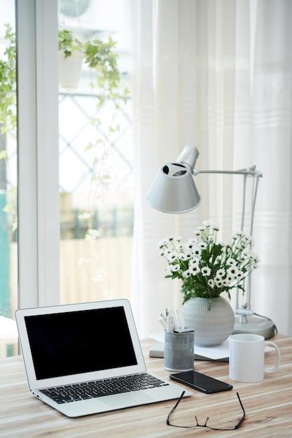 Portátil abierto con pantalla vacía, taza de café y jarrón con flores en el escritorio de la persona de negocios