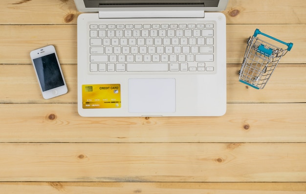 El portátil blanco con teléfono inteligente, tarjeta de crédito y el modelo de carrito de compras en la mesa de madera. compras de comercio electrónico.