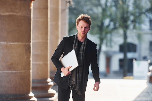 Con el portátil en las manos Elegante joven con gafas y ropa formal elegante al aire libre en la ciudad