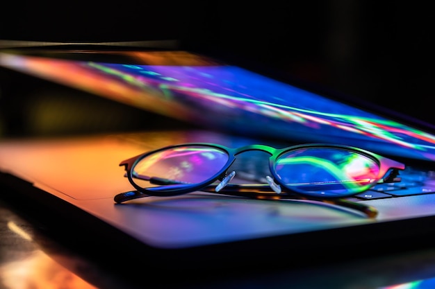 Un portátil medio cerrado en la oscuridad con un brillo colorido y gafas