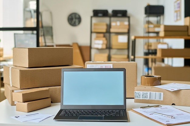 Portátil con pantalla en blanco entre documentos y cajas empaquetadas que contienen pedidos de clientes de tiendas en línea en la sala de almacenamiento