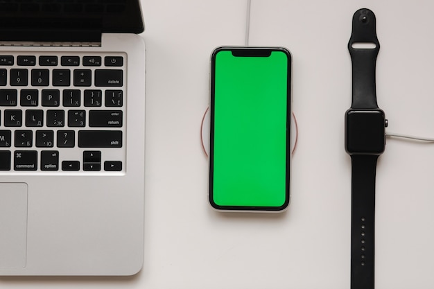 Portátil con reloj inteligente y carga de teléfono con cargador inalámbrico. Pantalla verde en el teléfono, indicador de carga en pantalla en el reloj. Vista superior. Lugar para el texto