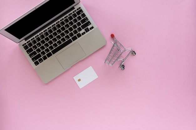 Portátil con tarjeta de crédito en blanco y mini carrito de compras sobre fondo rosa