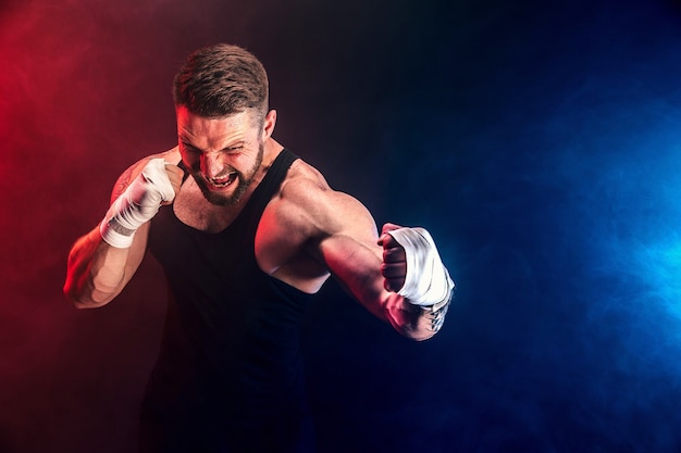 Porteiro barbudo e tatuado muay thai boxer em camiseta preta e luvas de boxe lutando na parede escura com fumaça. conceito de esporte.