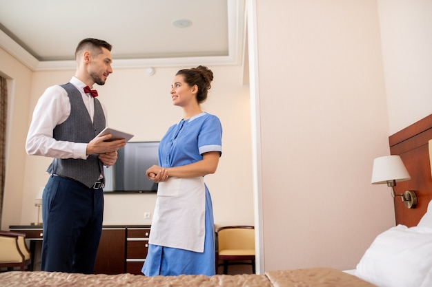Foto portero elegante joven con touchpad mirando a la criada bonita en uniforme durante la conversación en la habitación del hotel