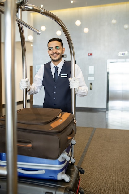 Portero de hotel joven en uniforme empujando el carro con el equipaje de los viajeros
