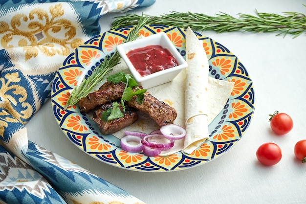 Portion Appetitlicher Beef Kebab, serviert mit Fladenbrot, Zwiebeln und roter Soße in einem gemusterten Teller auf einer weißen Tischdecke. Grill